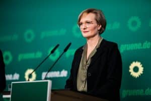 Franziska Brantner steht an einem Rednerpult vor einer grünen Wand mit dem Logo und der Schrift der Partei Bündnis 90/Die Grünen. Sie trägt eine Brille, eine dunkle Jacke und eine olivgrüne Bluse. Vor ihr befinden sich zwei Mikrofone.