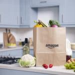 Eine braune Papiertüte mit dem Amazon-Logo steht auf einer modernen Küchenarbeitsplatte. Die Tüte ist mit frischem Obst und Gemüse gefüllt, darunter Brokkoli, Paprika und Spargel. Auf der Arbeitsplatte sind außerdem eine Flasche Olivenöl, eine Flasche Weißwein, Eier, Milch, Erdbeeren, Weintrauben und ein Blumenkohl zu sehen. Im Hintergrund befinden sich blaue Küchenschränke, ein Gasherd und verschiedene Küchenutensilien.