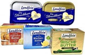 Ein Sortiment verschiedener Butterprodukte der Marke „Landfein“ in unterschiedlichen Verpackungen. Oben sind zwei blaue Plastikdosen „Die Streichbare“ zu sehen – eine mit Alpensalz, die andere ungesalzen. Darunter befinden sich drei Butterstücke in Papierverpackung: „Deutsche Süßrahmbutter“ in goldener Verpackung, „Deutsche Markenbutter“ in blauer Verpackung und „Original Irische Butter“ in goldener Verpackung mit grünem Aufdruck und einer Kuhillustration.