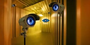 Futuristisch gestalteter „Big Brother“-Raum mit mehreren Überwachungskameras, deren Linsen wie blaue Augen aussehen. Im Hintergrund leuchtet das bekannte „Big Brother“-Logo mit stilisiertem Auge an einer gelb-orangenen Wand. Die Szenerie vermittelt eine dystopische