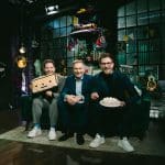 Christian Lindner sitzt zwischen den Zwillingen Benni und Dennis Wolter auf einem Sofa im Studio von „World Wide Wohnzimmer“. Benni hält ein Kissen mit Gesicht, Dennis eine Torte. Das Set ist bunt dekoriert mit Leuchtschildern, Retro-Objekten und Fanartikeln.