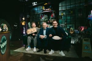 Christian Lindner sitzt zwischen den Zwillingen Benni und Dennis Wolter auf einem Sofa im Studio von „World Wide Wohnzimmer“. Benni hält ein Kissen mit Gesicht, Dennis eine Torte. Das Set ist bunt dekoriert mit Leuchtschildern, Retro-Objekten und Fanartikeln.