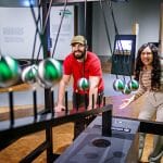Zwei begeisterte Besucher:innen interagieren mit einer physikalischen Pendel-Installation in der Ausstellung „Natur.Schau.Spiele.“ in der experimenta Heilbronn. Der Mann in einem roten T-Shirt und einer Kappe sowie die Frau mit langen Locken in einem gestreiften Pullover beobachten gespannt die schwingenden Metallkugeln. Im Hintergrund sind Informationstafeln zur Ausstellung zu sehen.