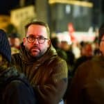 Paul Ronzheimer steht inmitten einer Demonstration am Abend. Der Journalist trägt eine Brille und eine dunkle Jacke, während er von weiteren Demonstranten umgeben ist. Im Hintergrund sind rote Fahnen und Lichter der Stadt zu sehen.
