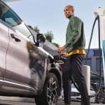 Ein Mann lädt sein Elektroauto an einer Shell Recharge Schnellladestation auf. Er steht neben dem Fahrzeug, hält das Ladekabel und blickt in die Kamera. Die Ladesäule ist mit einem Shell-Logo und Informationen zur Ladeleistung von 360 kW versehen. Im Hintergrund sind weitere Fahrzeuge und ein Gebäude zu sehen.