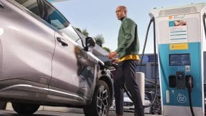 Ein Mann lädt sein Elektroauto an einer Shell Recharge Schnellladestation auf. Er steht neben dem Fahrzeug, hält das Ladekabel und blickt in die Kamera. Die Ladesäule ist mit einem Shell-Logo und Informationen zur Ladeleistung von 360 kW versehen. Im Hintergrund sind weitere Fahrzeuge und ein Gebäude zu sehen.