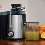 Ein moderner Zentrifugalentsafter aus Edelstahl mit einem vollen Saftbehälter steht auf einer Küchenarbeitsplatte. Daneben liegen frisch aufgeschnittene Orangen, während im Hintergrund ein Korb mit weiteren Zitrusfrüchten und eine Vase mit Blumen zu sehen sind.