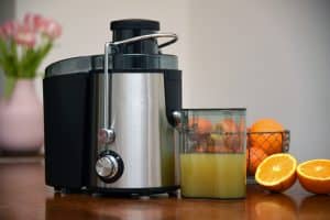 Ein moderner Zentrifugalentsafter aus Edelstahl mit einem vollen Saftbehälter steht auf einer Küchenarbeitsplatte. Daneben liegen frisch aufgeschnittene Orangen, während im Hintergrund ein Korb mit weiteren Zitrusfrüchten und eine Vase mit Blumen zu sehen sind.