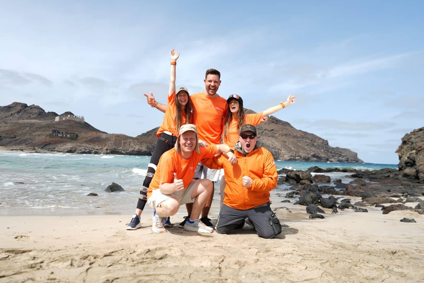 Fünf Mitglieder von Team Orange aus der Serie „Mission Unknown“ stehen jubelnd am Strand, gekleidet in orangefarbene Shirts. Hinter ihnen das raue Meer und felsige Küstenlandschaft – Symbol für die überstandene Herausforderung der Atlantiküberquerung.







