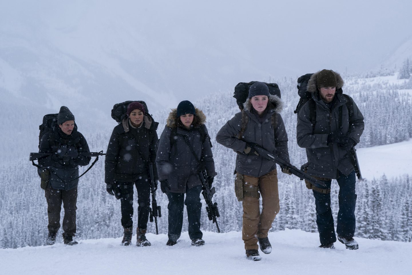 Eine Gruppe von fünf bewaffneten Menschen, darunter Ellie (Bella Ramsey) und Abby (Kaitlyn Dever), wandert durch eine verschneite Berglandschaft. Sie tragen schwere Winterkleidung und Rucksäcke, während Schneeflocken um sie herumfallen. Das Bild stammt aus der zweiten Staffel der HBO-Serie „The Last of Us“.
