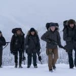 Eine Gruppe von fünf bewaffneten Menschen, darunter Ellie (Bella Ramsey) und Abby (Kaitlyn Dever), wandert durch eine verschneite Berglandschaft. Sie tragen schwere Winterkleidung und Rucksäcke, während Schneeflocken um sie herumfallen. Das Bild stammt aus der zweiten Staffel der HBO-Serie „The Last of Us“.