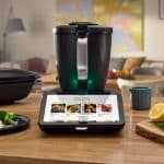 Der neue Thermomix TM7 steht auf einer Küchenarbeitsplatte. Der smarte Küchenhelfer hat ein modernes Design mit einem großen Touchscreen, auf dem Rezeptvorschläge angezeigt werden. Im Vordergrund liegen frische Zutaten wie Zitronen und Dill auf einem Holzbrett, daneben steht ein Teller mit Lachs, Reis und Brokkoli. Im Hintergrund ist ein gemütlich eingerichtetes Wohnzimmer zu sehen.
