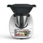 Vorwerk Thermomix TM6 – eine moderne Küchenmaschine mit Touchscreen, Edelstahl-Mixtopf und schwarzen Aufsätzen, darunter ein Varoma-Dampfgarer. Das Display zeigt eine Rezeptauswahl.