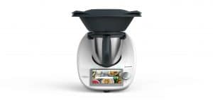 Vorwerk Thermomix TM6 – eine moderne Küchenmaschine mit Touchscreen, Edelstahl-Mixtopf und schwarzen Aufsätzen, darunter ein Varoma-Dampfgarer. Das Display zeigt eine Rezeptauswahl.