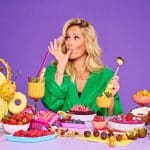 Helene Fischer sitzt vor einem bunt gedeckten Frühstückstisch mit verschiedenen Wholey-Produkten, tropischen Früchten und Smoothies. Sie trägt einen grünen Bademantel, hält einen goldenen Löffel in der Hand und leckt sich genüsslich den Daumen. Der Hintergrund ist lila, und die Szene wirkt farbenfroh