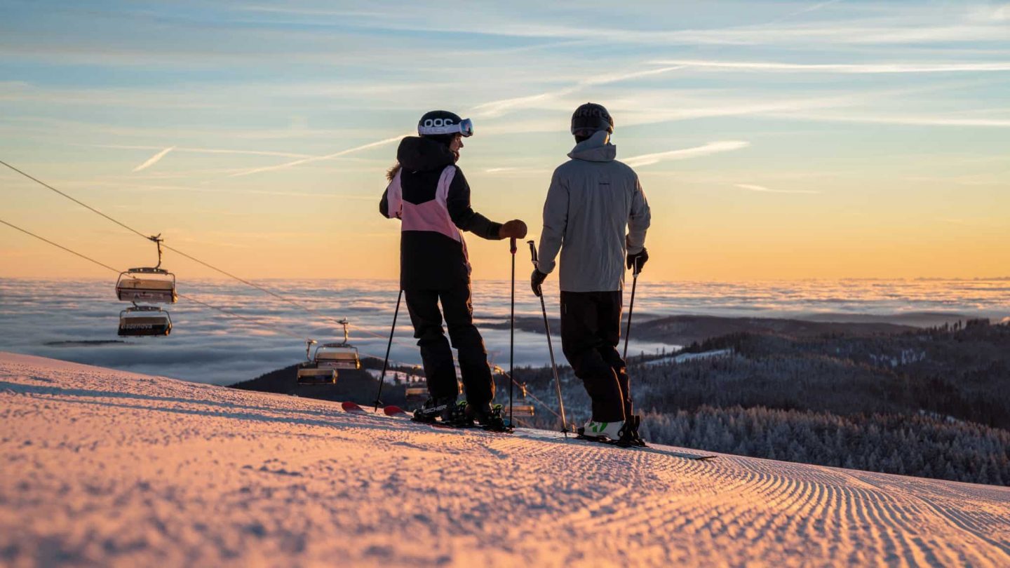 Zwei Skifahrer stehen auf einer frisch präparierten Piste am Feldberg und blicken bei Sonnenuntergang über ein beeindruckendes Nebelmeer. Im Hintergrund schweben Sessellifte über die winterliche Landschaft.