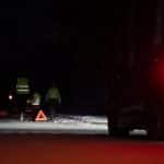 Eine Familie mit Warnwesten steht bei Dunkelheit auf einer verschneiten Straße neben einem Warndreieck, während ein Auto mit eingeschalteten Rücklichtern vorbeifährt.