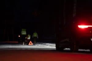 Eine Familie mit Warnwesten steht bei Dunkelheit auf einer verschneiten Straße neben einem Warndreieck, während ein Auto mit eingeschalteten Rücklichtern vorbeifährt.