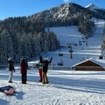 Auf dem Bild ist ein malerisches Skigebiet in den Alpen zu sehen. Der Himmel ist strahlend blau, die Berge und dichten Nadelwälder sind mit Schnee bedeckt. Eine Gruppe von Skifahrern und Snowboardern steht auf einer Aussichtsplattform und scheint sich zu unterhalten. Im Vordergrund liegen Snowboards und Skier auf dem Schnee. Ein Sessellift führt die Piste hinauf, auf der mehrere Wintersportler unterwegs sind. Die Szenerie wirkt idyllisch und lädt zum Skifahren ein.