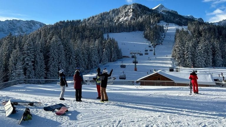 Auf dem Bild ist ein malerisches Skigebiet in den Alpen zu sehen. Der Himmel ist strahlend blau, die Berge und dichten Nadelwälder sind mit Schnee bedeckt. Eine Gruppe von Skifahrern und Snowboardern steht auf einer Aussichtsplattform und scheint sich zu unterhalten. Im Vordergrund liegen Snowboards und Skier auf dem Schnee. Ein Sessellift führt die Piste hinauf, auf der mehrere Wintersportler unterwegs sind. Die Szenerie wirkt idyllisch und lädt zum Skifahren ein.