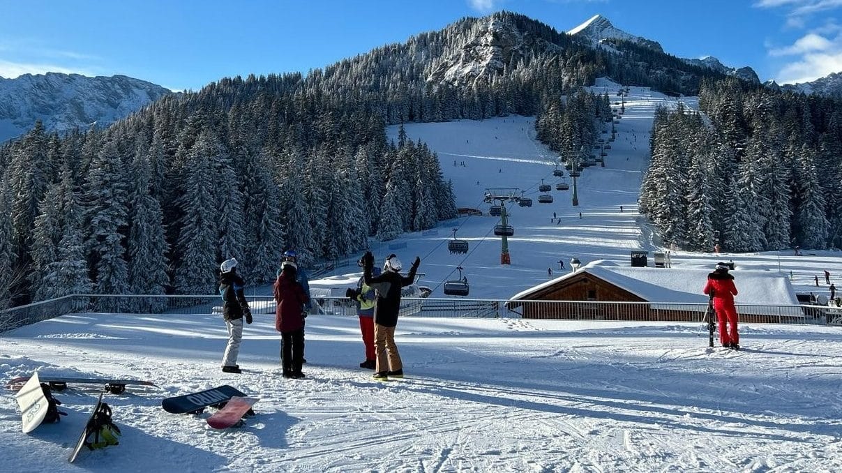 Auf dem Bild ist ein malerisches Skigebiet in den Alpen zu sehen. Der Himmel ist strahlend blau, die Berge und dichten Nadelwälder sind mit Schnee bedeckt. Eine Gruppe von Skifahrern und Snowboardern steht auf einer Aussichtsplattform und scheint sich zu unterhalten. Im Vordergrund liegen Snowboards und Skier auf dem Schnee. Ein Sessellift führt die Piste hinauf, auf der mehrere Wintersportler unterwegs sind. Die Szenerie wirkt idyllisch und lädt zum Skifahren ein.