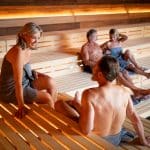 Menschen entspannen in einer modernen Sauna mit hölzernen Sitzbänken und warmer Beleuchtung. Zwei Personen sitzen im Vordergrund im Gespräch, während ein weiteres Paar im Hintergrund liegt und sich ausruht.