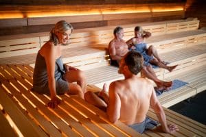 Menschen entspannen in einer modernen Sauna mit hölzernen Sitzbänken und warmer Beleuchtung. Zwei Personen sitzen im Vordergrund im Gespräch, während ein weiteres Paar im Hintergrund liegt und sich ausruht.
