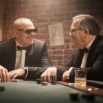 Stefan Raab und Elton im Anzug am Pokertisch: Die beiden TV-Stars lachen und unterhalten sich in einer stilvollen Poker-Runde. Vor ihnen liegen Karten, Chips und Gläser, während eine Backsteinwand im Hintergrund das stimmungsvolle Setting abrundet
