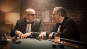 Stefan Raab und Elton im Anzug am Pokertisch: Die beiden TV-Stars lachen und unterhalten sich in einer stilvollen Poker-Runde. Vor ihnen liegen Karten, Chips und Gläser, während eine Backsteinwand im Hintergrund das stimmungsvolle Setting abrundet