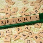 Ein Scrabble-Spielbrett mit Holzbuchstaben, die das Wort „Zeugnis“ bilden. Die Buchstaben liegen auf einem grünen Halter, während weitere verstreute Buchstabensteine auf einem strukturierten, grün-weißen Untergrund verteilt sind.