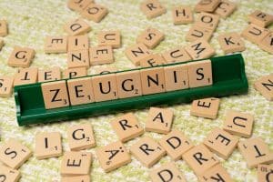 Ein Scrabble-Spielbrett mit Holzbuchstaben, die das Wort „Zeugnis“ bilden. Die Buchstaben liegen auf einem grünen Halter, während weitere verstreute Buchstabensteine auf einem strukturierten, grün-weißen Untergrund verteilt sind.