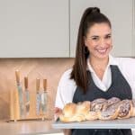 Saliha Özcan, bekannt als Sally, steht in einer modernen Küche und hält ein Backblech mit frisch gebackenem Hefezopf. Sie trägt eine weiße Bluse und eine dunkle Schürze und lächelt in die Kamera. Im Hintergrund sind Küchenutensilien, eine Küchenmaschine und eine stilvolle Kücheneinrichtung zu sehen.