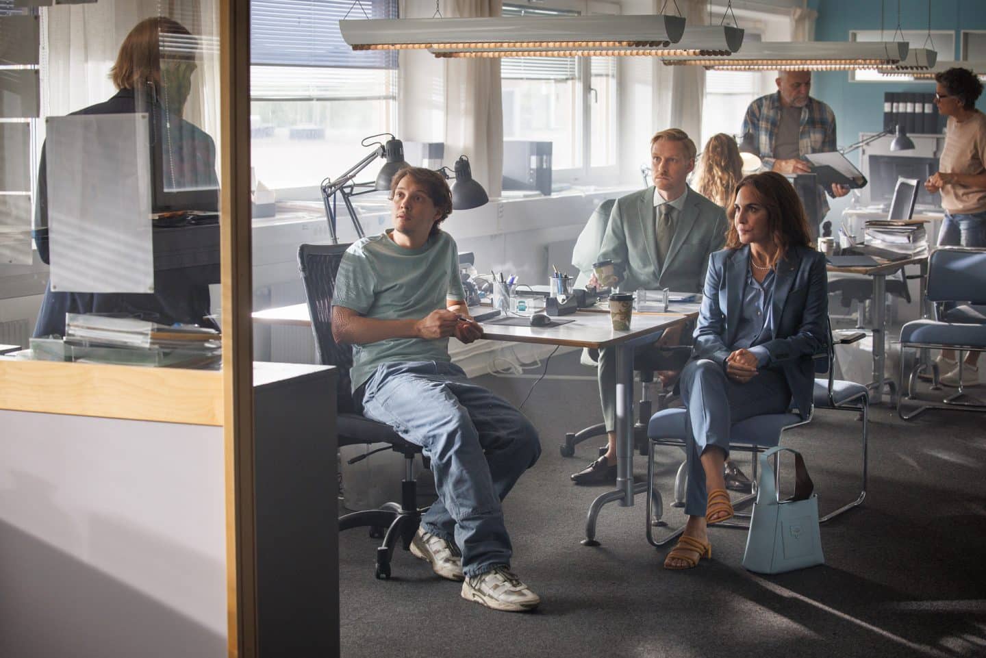 "Mord im Mittsommer - Madeleine": Im Büro: Ermittler Welpe (Julius Fleischanderl), Staatsanwältin Nora Linde (Alexandra Rapaport) und ihr Mitarbeiter Pär (Anton Lundqvist) sitzen im Polizeirevier um einen Schreibtisch herum. Sie blicken in die Richtung von Polizeichef Stenmark (Gustaf Hammarsten), der ihnen gegenübersteht. Stenmark ist nur von hinten zu sehen. Im Hintergrund arbeiten drei Kollegen (Komparsen).
