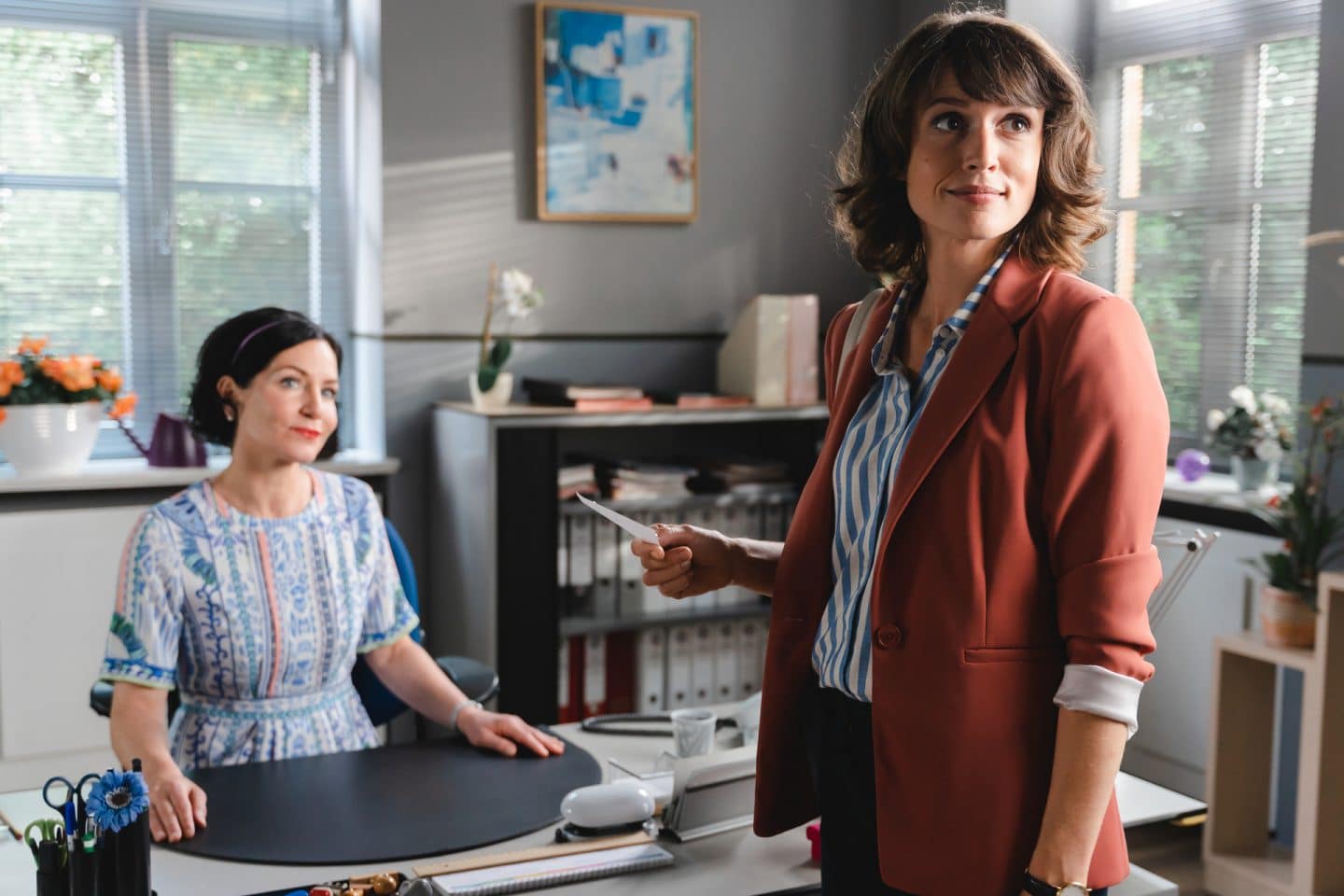 "Die Rosenheim-Cops - Ein überraschendes Wiedersehen": Miriam Stockl (Marisa Burger) sitzt an ihrem Schreibtisch im Kommissariat und betrachtet lächelnd Kommissarin Andresen (Sophie Melbinger), die mit einem Zettel in der Hand vor ihr steht. Im Hintergrund ist ein Aktenschrank zu sehen.
