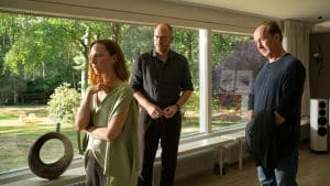 "Wendland - Stiller und der Teufelssauger": Jakob Stiller (Ulrich Noethen), Oda Nolde (Katharina Schüttler) und Jan Nolde (Johann von Bülow) stehen zusammen in einem Wohnzimmer. Oda Nolde steht mit dem Rücken zu Jakob Stiller und Jan Nolde. Hinter ihnen ist der Garten durch eine Fensterfront zu sehen.