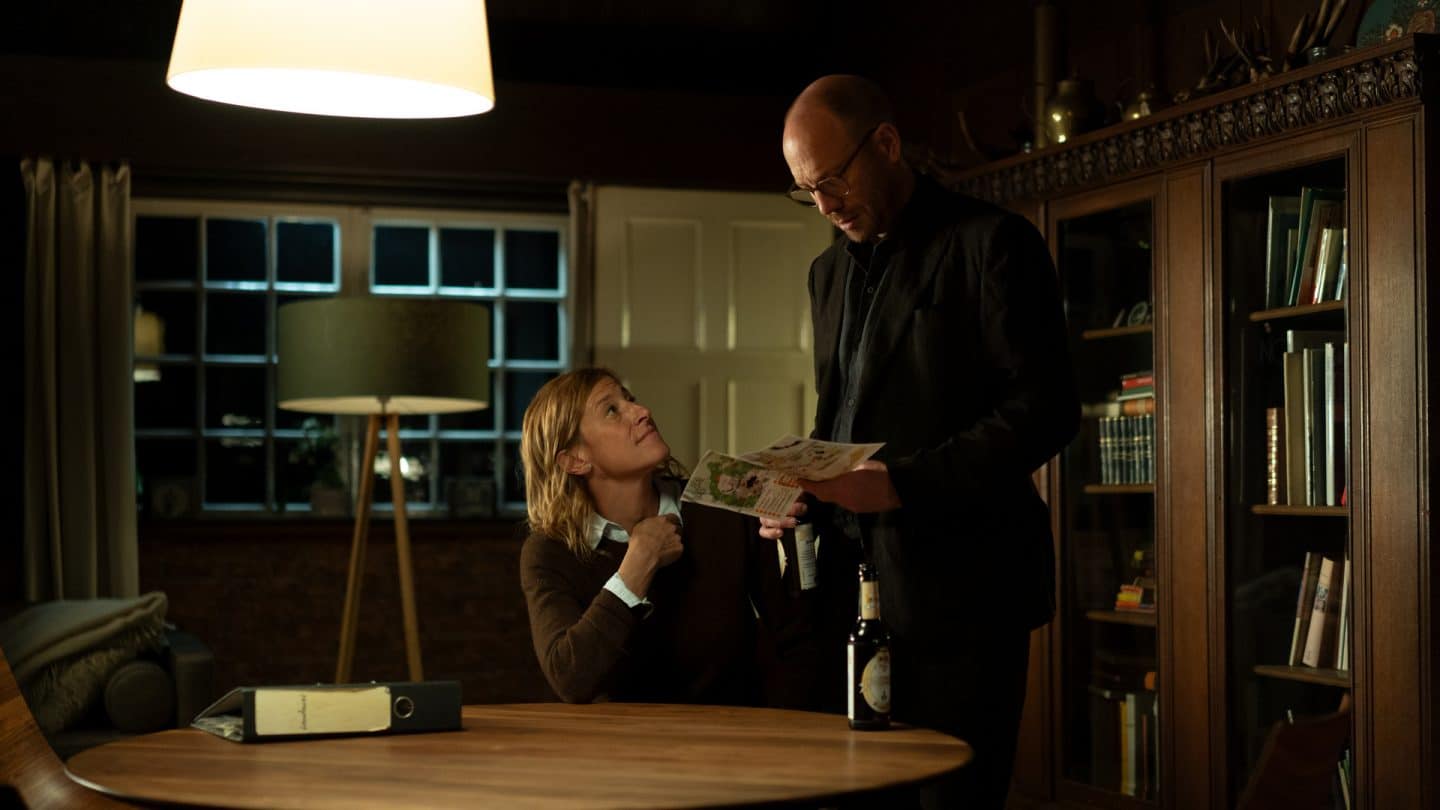 "Wendland - Stiller und der Teufelssauger": Abends im Lampenschein: Silke Landauer (Helene Grass) sitzt im Wohnzimmer an einem Tisch und zeigt Jan Nolde (Johann von Bülow) einen Zeitungsausschnitt.
