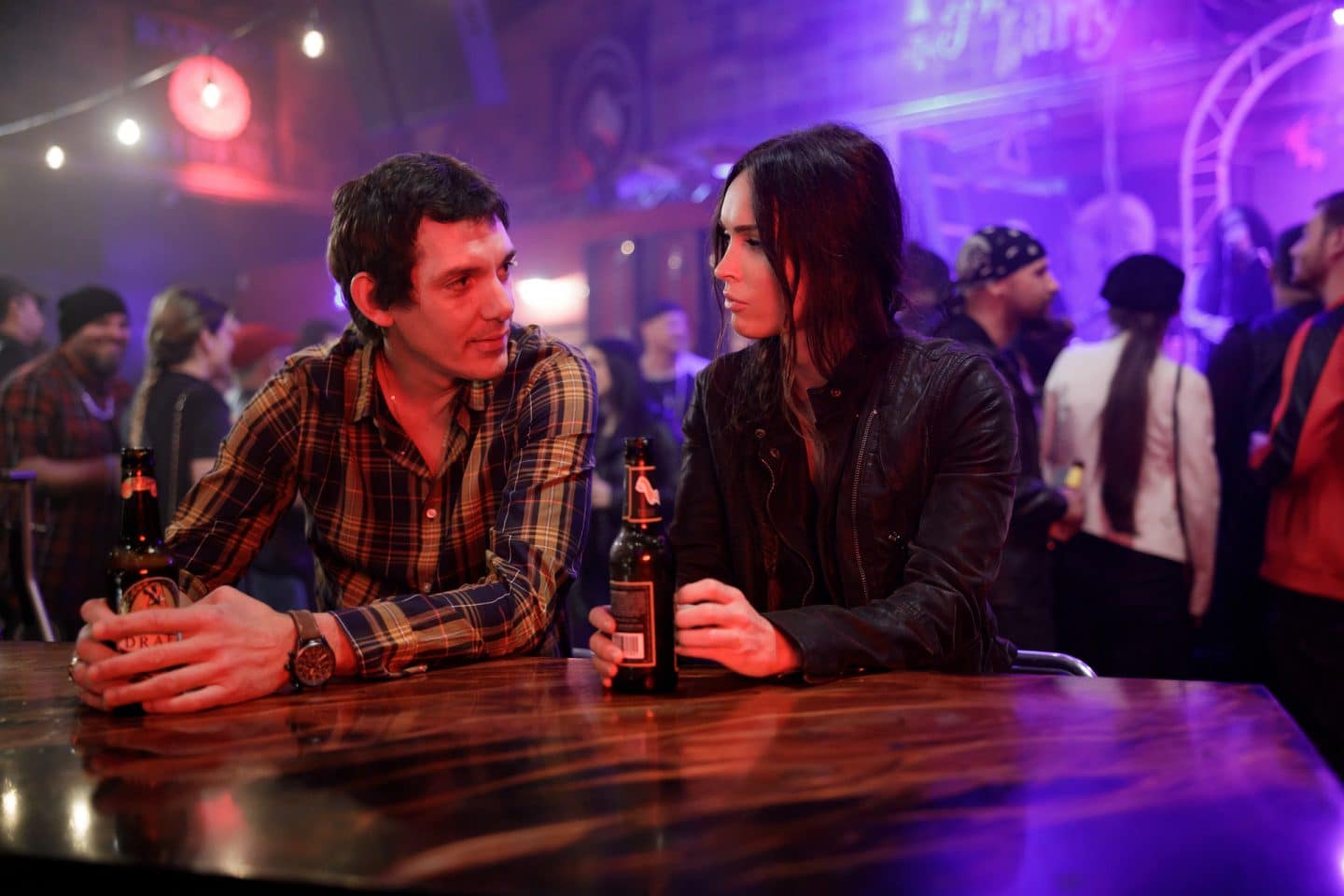 "Die Spur des Killers – Midnight in the Switchgrass": Rebecca Lombardi (Megan Fox) wird in einer Bar an einem Einzeltisch von Peter (Lukas Haas) angesprochen. Beide halten eine Bierflasche in der Hand. Im Hintergrund stehen und sitzen weitere Besucher der Bar.
