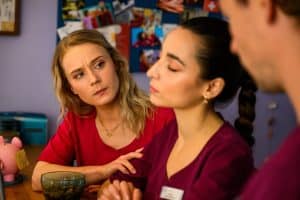 "Bettys Diagnose - Verfahren": Mia (Antonia Döring) und Lukas (Niklas Löffler) beobachten Ava (Rona Özkan), die mit geschlossenen Augen zwischen ihnen sitzt. Neben Mia steht ein rosafarbenes Sparschwein auf einem Tresen. Im Hintergrund hängt eine Pinnwand mit verschiedenen Bildern.