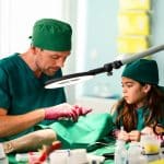 "Bettys Diagnose - Liebe hat viele Gesichter": Dr. Walden (Constantin Lücke) schaut durch eine OP-Lampe und erklärt der kleinen Jonna (Soraya Maria Efe), wie eine OP funktioniert. Im Vordergrund sind verschiedene OP-Utensilien zu erkennen.