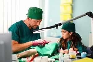 "Bettys Diagnose - Liebe hat viele Gesichter": Dr. Walden (Constantin Lücke) schaut durch eine OP-Lampe und erklärt der kleinen Jonna (Soraya Maria Efe), wie eine OP funktioniert. Im Vordergrund sind verschiedene OP-Utensilien zu erkennen.