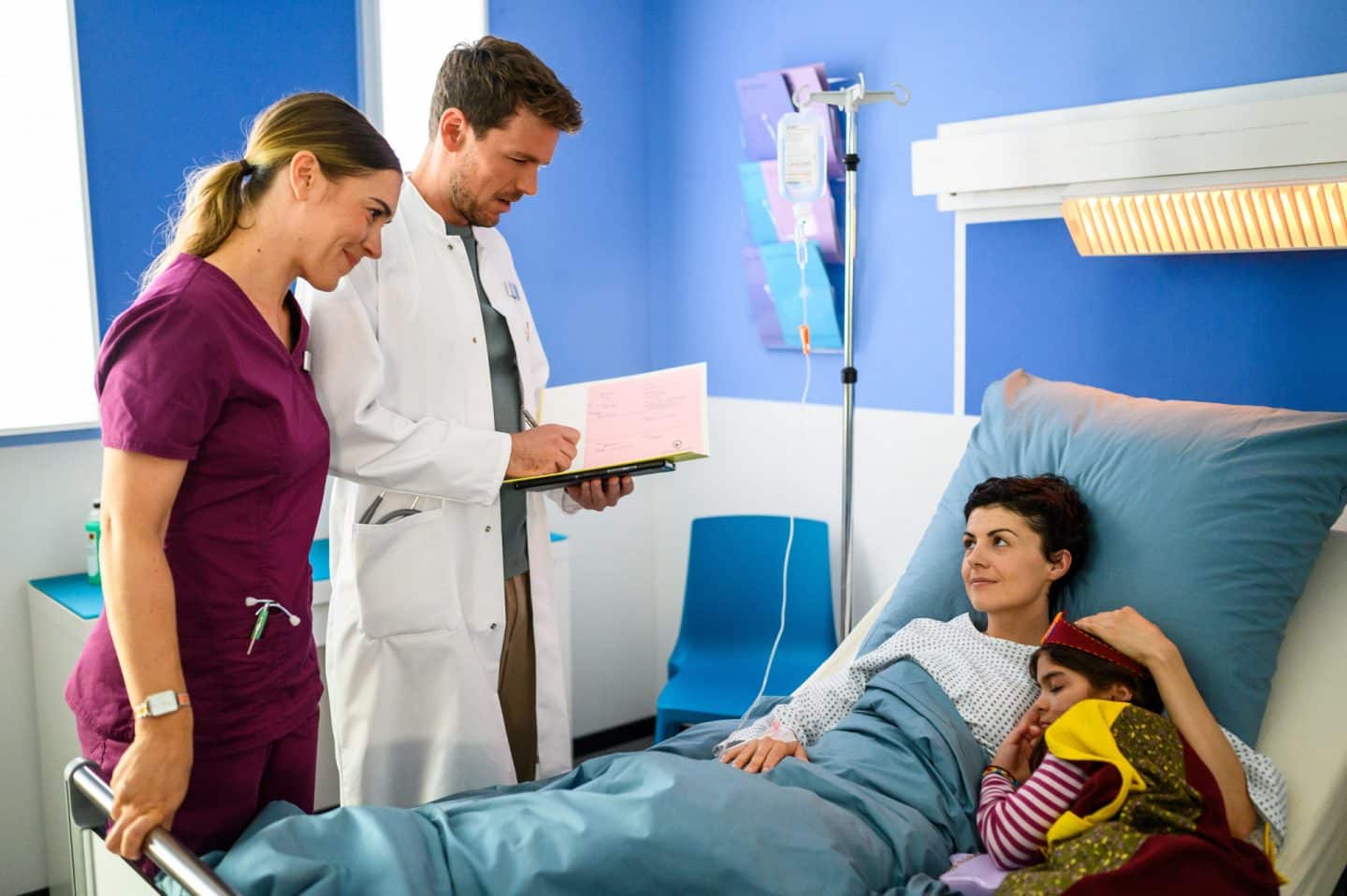 "Bettys Diagnose - Liebe hat viele Gesichter": Betty (Henrike Hahn) und Dr. Walden (Constantin Lücke) stehen am Krankenbett von Iris Herdinger (Liza Tzschirner), die ihre Tochter Jonna (Soraya Maria Efe) im Arm hält. Dr. Walden hält die Patientenakte in den Händen.
