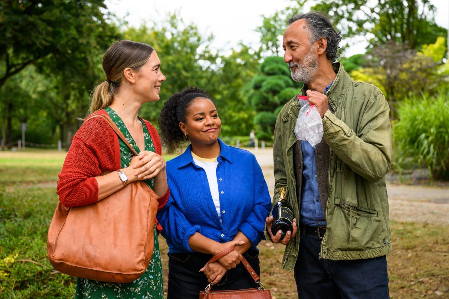 "Bettys Diagnose - Liebe hat viele Gesichter": Betty (Henrike Hahn) und Zoe (Catherine Chikosi) unterhalten sich im Park mit Safi (Ercan Durmaz), der eine Flasche Sekt und eine Plastiktüte in den Händen hält. Betty und Zoe halten dabei ihre Handtaschen in den Händen.
