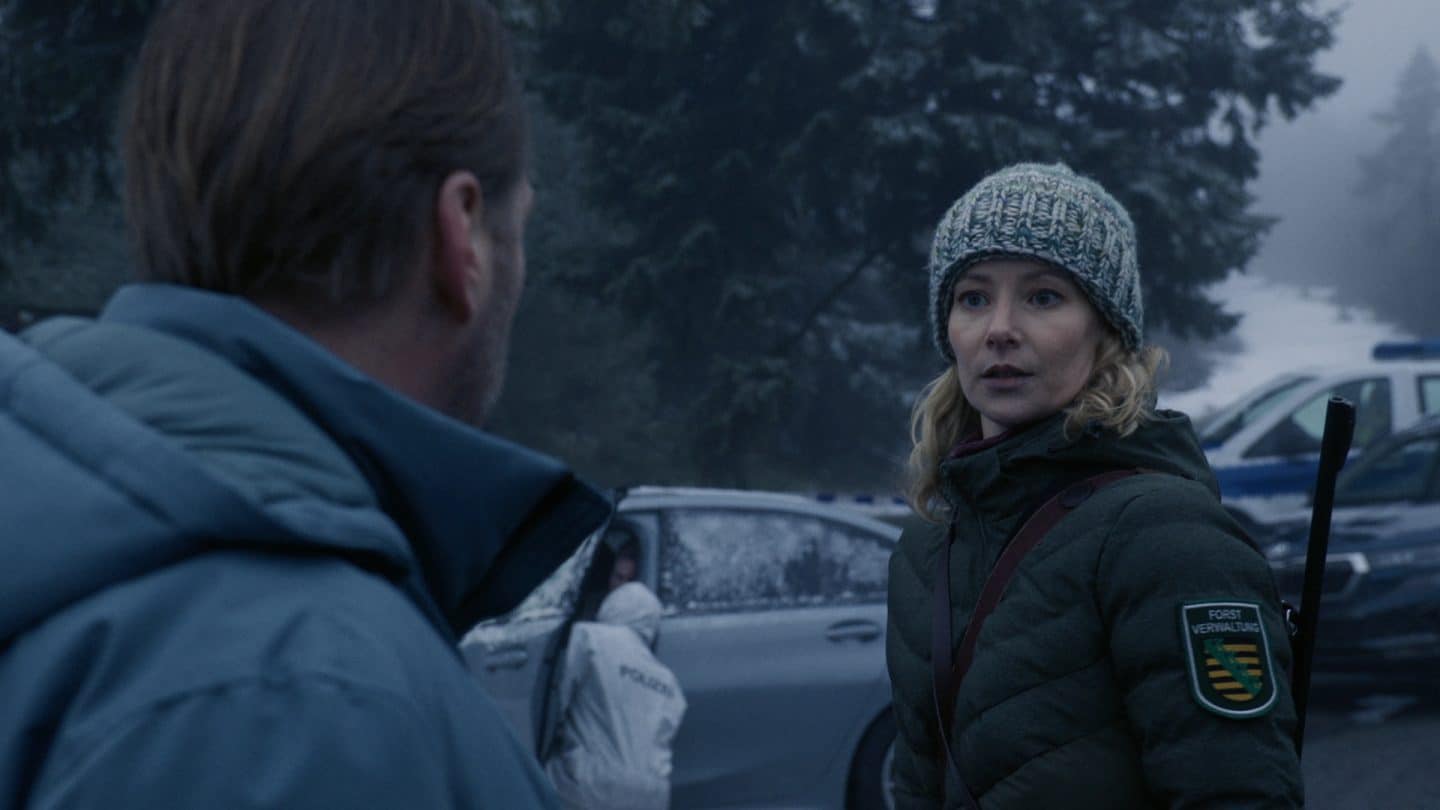 "Erzgebirgskrimi - Wintermord": Försterin Saskia Bergelt (Teresa Weißbach) blickt besorgt zu Hauptkommissar Robert Winkler (Kai Scheve). Im Hintergrund stehen mehrere Autos und jemand von der Spurensicherung arbeitet an dem Auto.