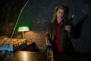 "Der Staatsanwalt - Verfeindet": Anita Brehm (Leslie Malton) steht in einem dunklen Kellerraum und sieht auf eine geöffnete Weinflasche, die sie in ihrer linken Hand hält. In ihrer rechten Hand hält sie einen Korken. Vor und neben ihr ist ein Tisch, auf dem ein volles Glas, ein Tablett mit weiteren Gläsern und eine kleine grüne Stehlampe stehen.