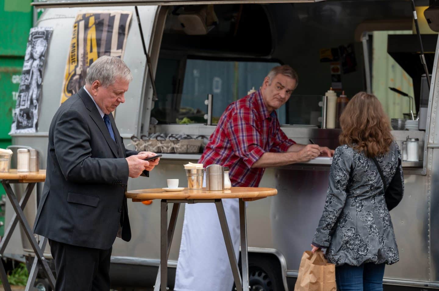 "Der Staatsanwalt - Tödliche Nähe": Bernd Reuther (Rainer Hunold) steht an einem runden Stehtisch neben einem Imbisswagen. Er schaut konzentriert auf sein Smartphone. Auf dem Stehtisch steht unter anderem eine Espressotasse. Stefan Schiller (Heinrich Schafmeister) steht im Hintergrund an der Theke des Imbisswagens und schaut zu Bernd Reuther hinüber. Schiller hält einen Stift in der Hand, ihm gegenüber steht eine Frau (Komparsin), die eine Papiertüte in der linken Hand hält.
