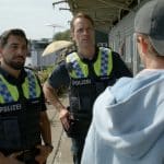 "Notruf Hafenkante - Kein Platz auf dieser Welt": Die Polizisten Malik Hakim (Zejhun Demirov) und Mattes Seeler (Matthias Schloo) stehen nebeneinander vor Wohncontainern. Beide blicken zu Colin (Aris Wolf), der seitlich vor ihnen steht. Man sieht ihn von hinten.