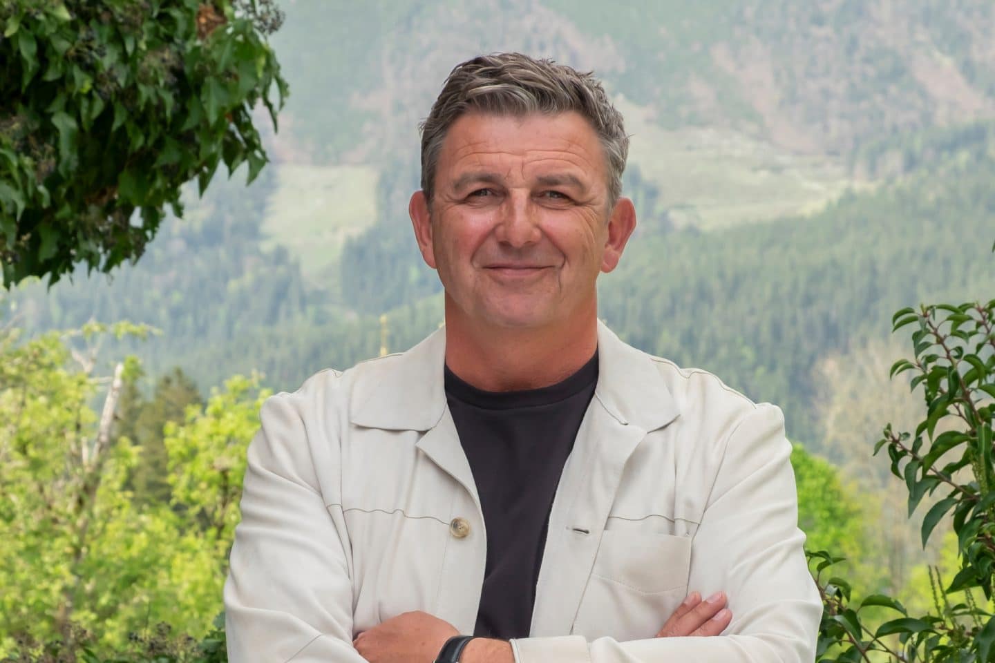 "Der Bergdoktor": Vor einer grünen Bergkulisse steht, die Arme vor der Brust verschränkt, Dr. Martin Gruber (Hans Sigl) und lächelt in die Kamera.