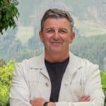 "Der Bergdoktor": Vor einer grünen Bergkulisse steht, die Arme vor der Brust verschränkt, Dr. Martin Gruber (Hans Sigl) und lächelt in die Kamera.