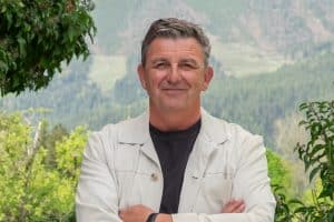 "Der Bergdoktor": Vor einer grünen Bergkulisse steht, die Arme vor der Brust verschränkt, Dr. Martin Gruber (Hans Sigl) und lächelt in die Kamera.