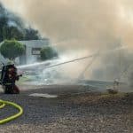 "Marie fängt Feuer - Vergeben und Vergessen": Marie (Christine Eixenberger) kniet zusammen mit einem Kollegen der Feuerwehr auf dem Boden des Campingplatzes Beide zielen mit Feuerwehrschläuchen auf brennende Wohnwagen, die sich hinter einer Rauchwolke befinden.